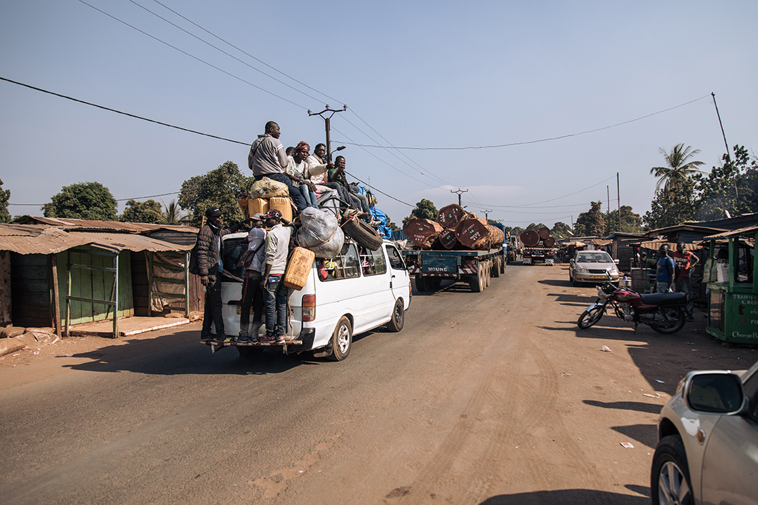 People sit on top of a white van as it drives through a town.