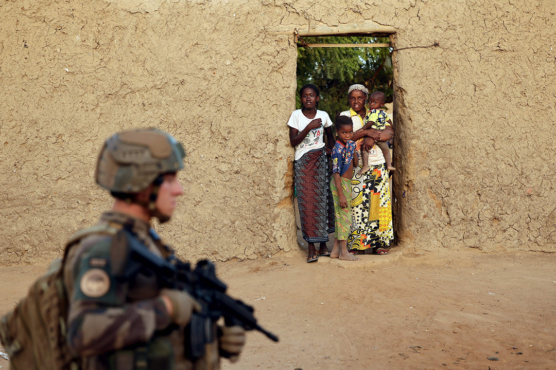 A soldier patrols in front of a building where a woman and three children stand.