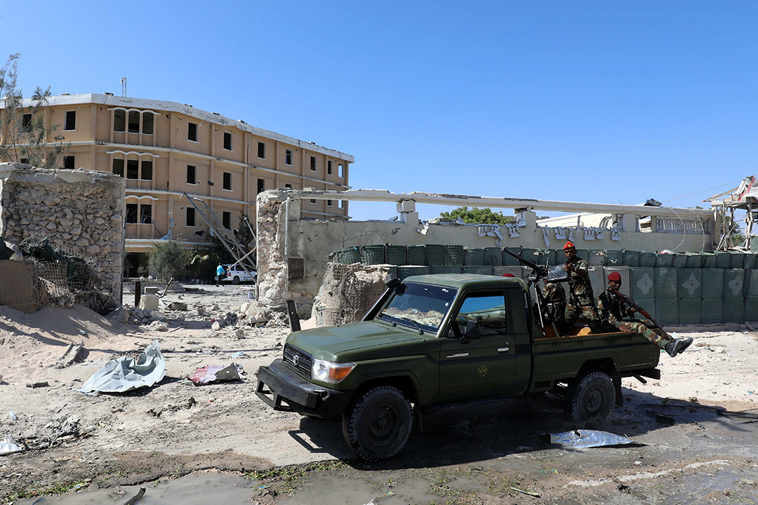 Men wearing military fatigues sit in the back of a green pickup truck outsude a building with a demolished security perimeter.