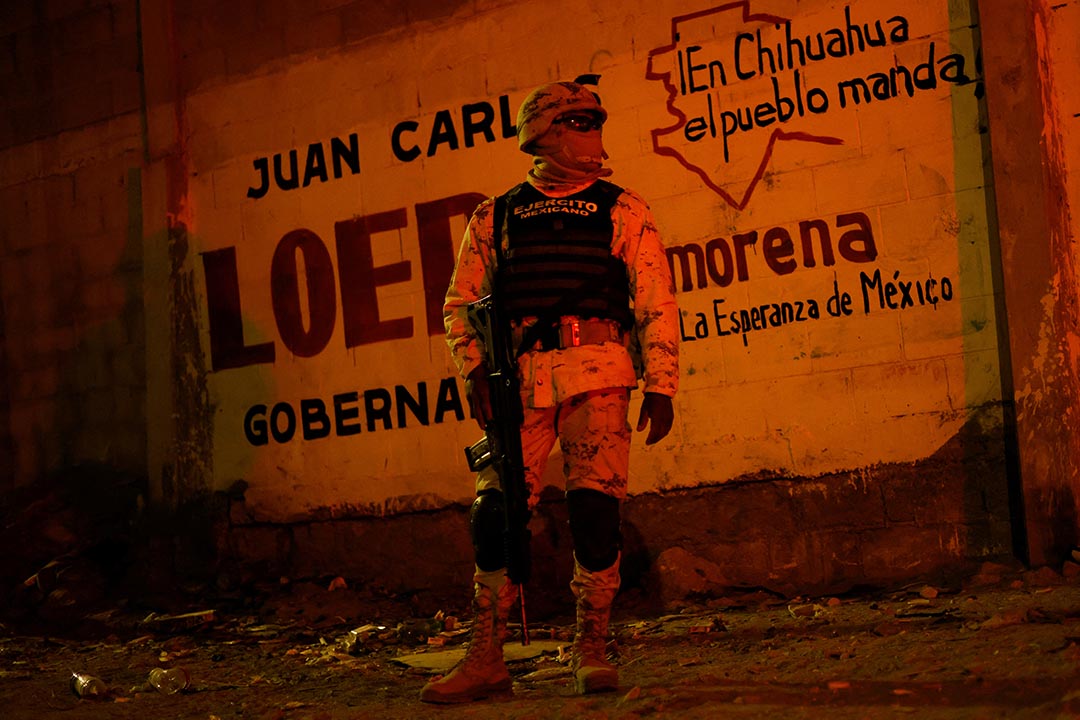 A person in military fatigues stands against a wall facing the camera.