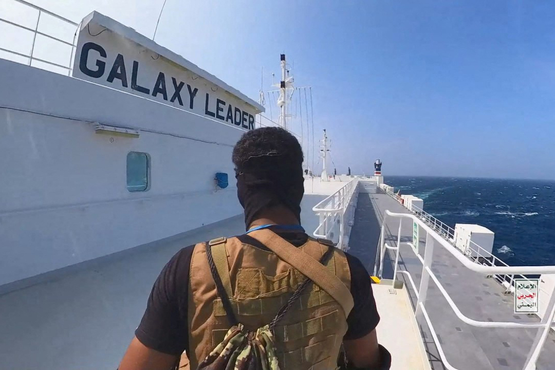 A man wearing military gear faces away from the camera while walking on a white cargo ship.