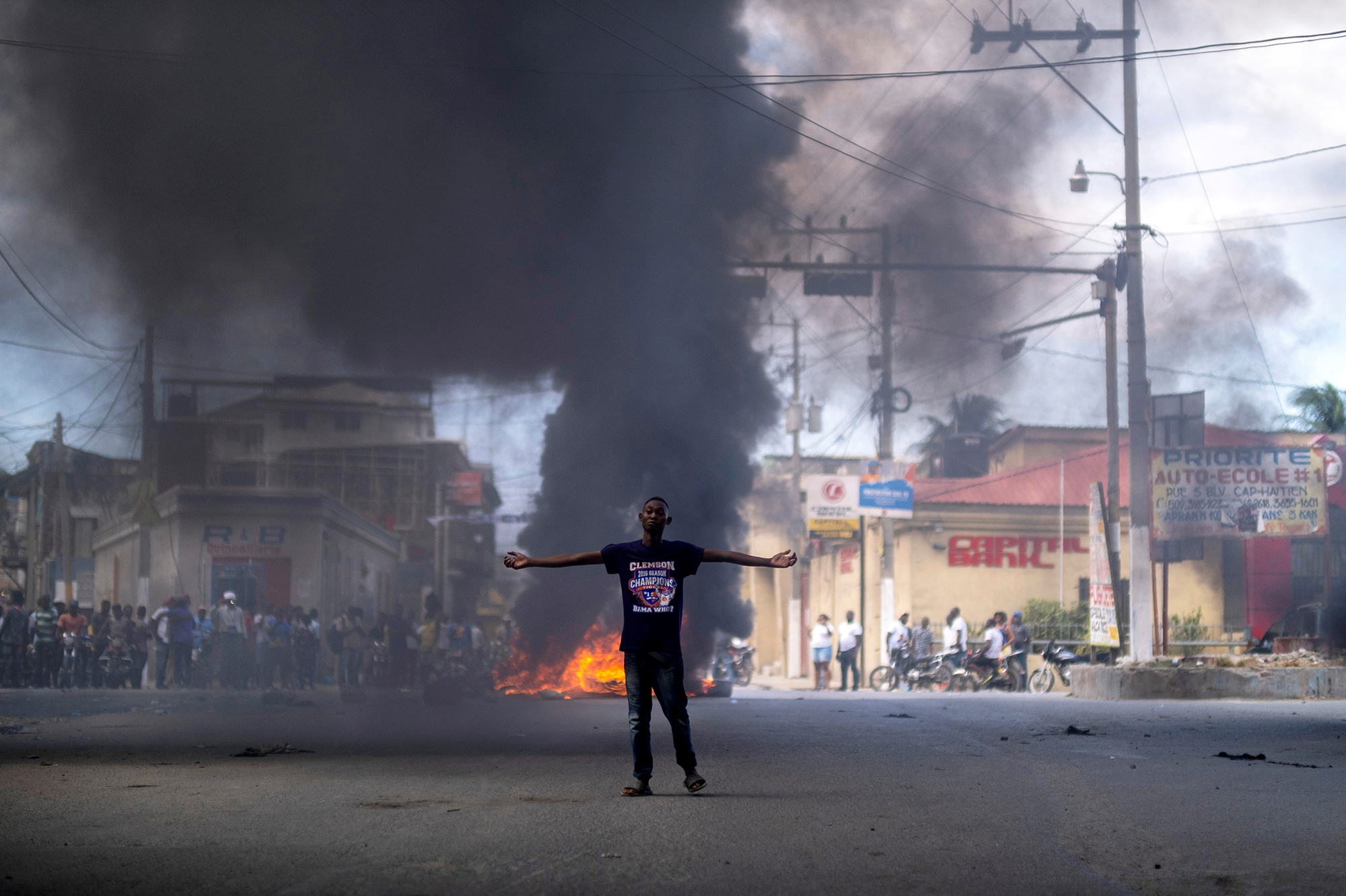 A young man wearing a black t-shirt and jeans stands in front a burning barricade in a street.