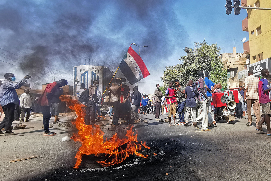 Protestors stand behind a burning tire in a street, one person holds a flag.