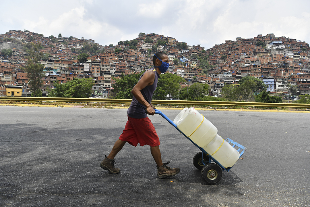 A man pushes a cart carrying water containers on a road while crowded houses on a mountain are in the distance.