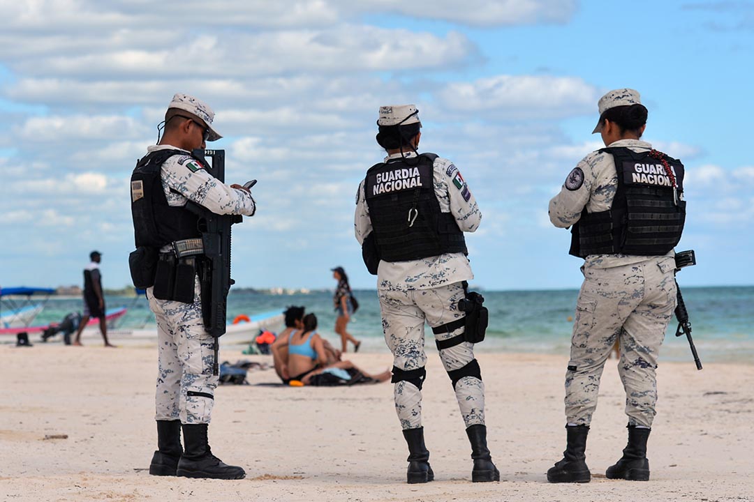 Three peple stand facing away from the camera wearing military uniforms on a white beach with a blue sky.