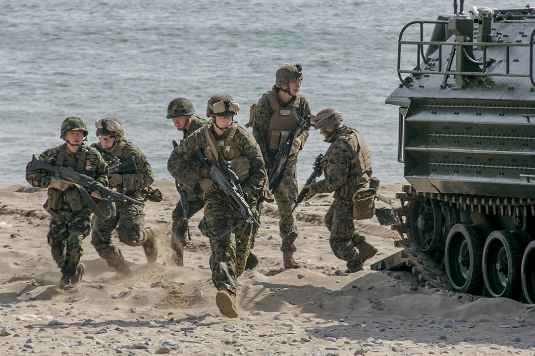 Six people wearing military uniforms run on the beach next to a tank.