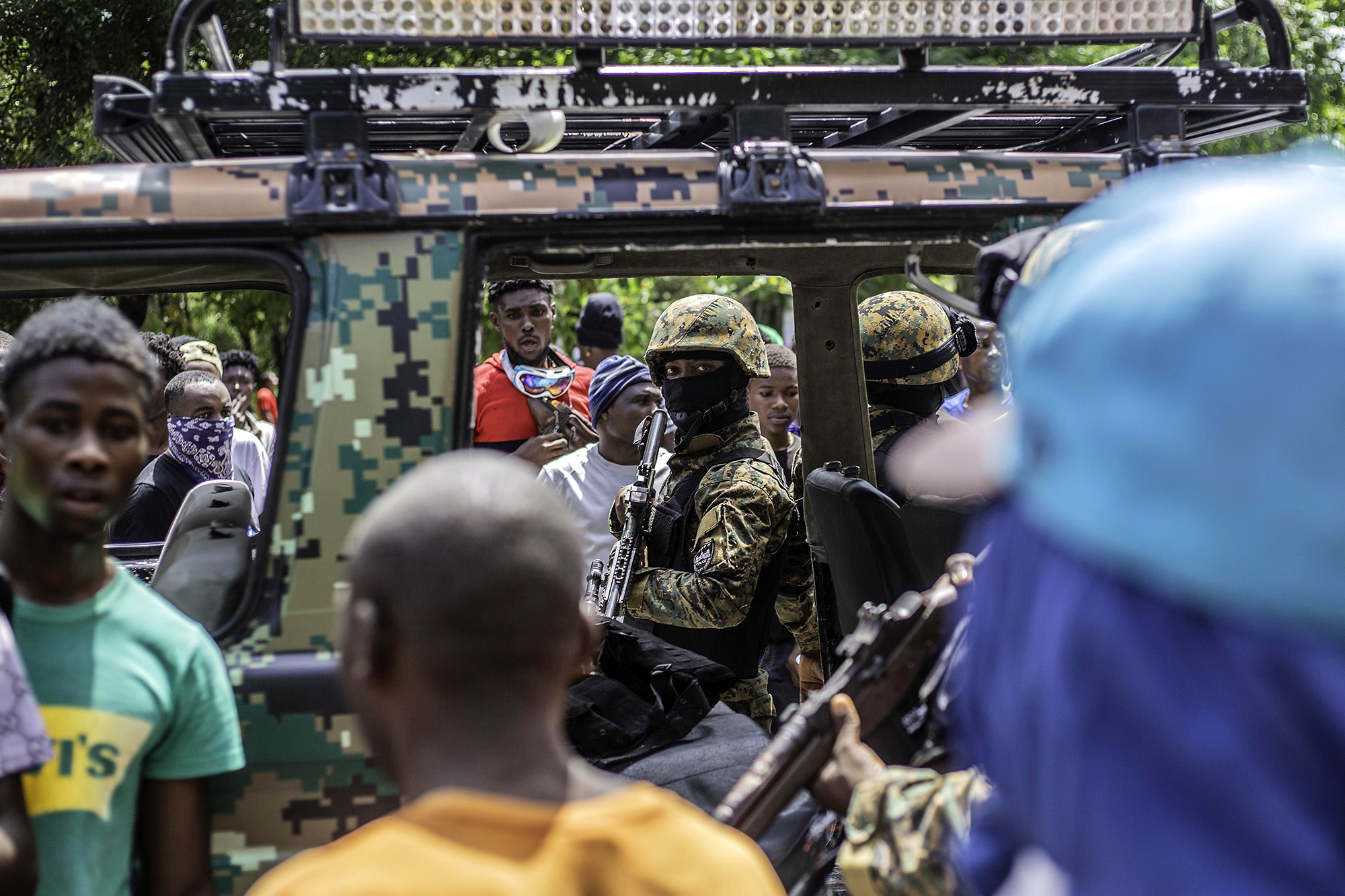 A soldier sits in a military vehicle while looking at civilians that surround the car.