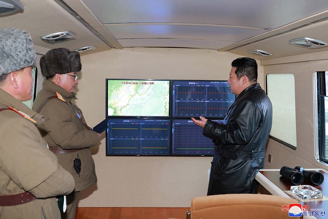 Leader Kim Jong Un wears a black leather jacket while standing in front of screens as two officials wear heavy coats and hats stand next to him.