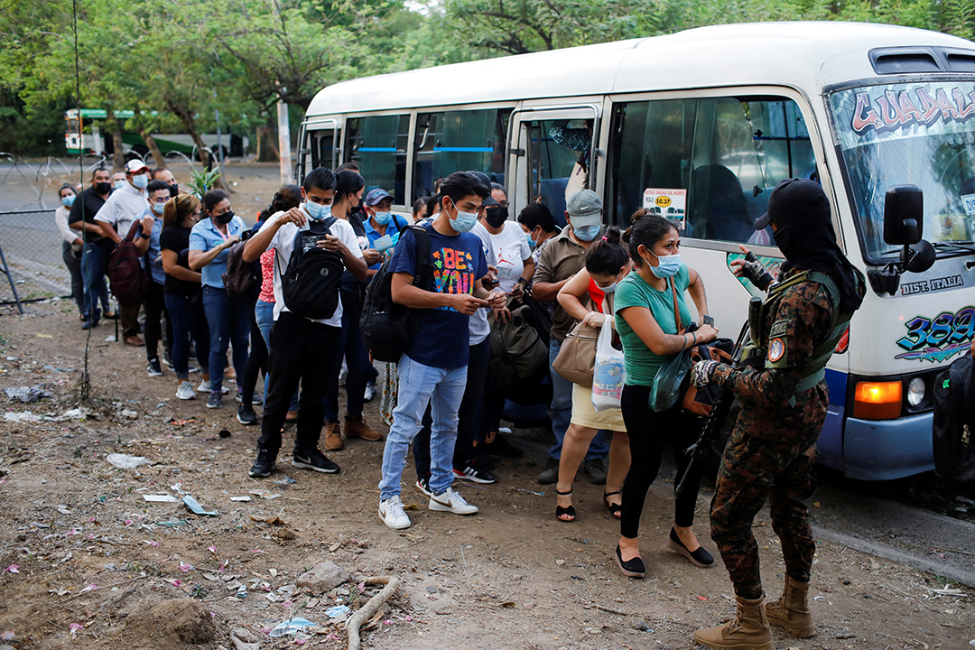 A group of men and women line up beside a white bus as they are checked by a police officer.