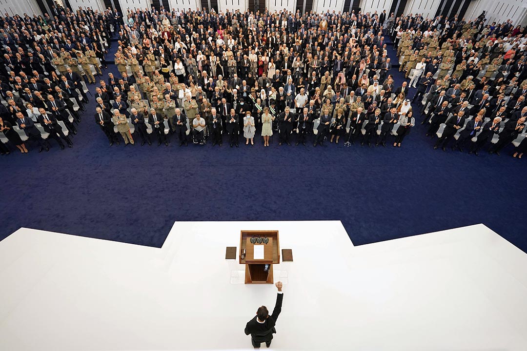 Syrian president stands behind a wooden podium on a white stage facing a crowd of people.