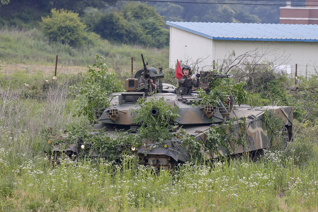 Two members in the military stand inside a tank near a wooded area.