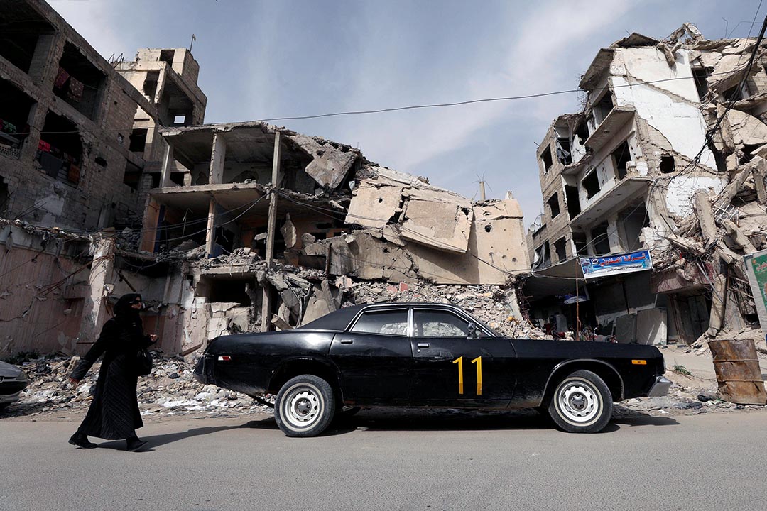 A woman in a black veil walks past a black vintage car parked in front of a demolished building.