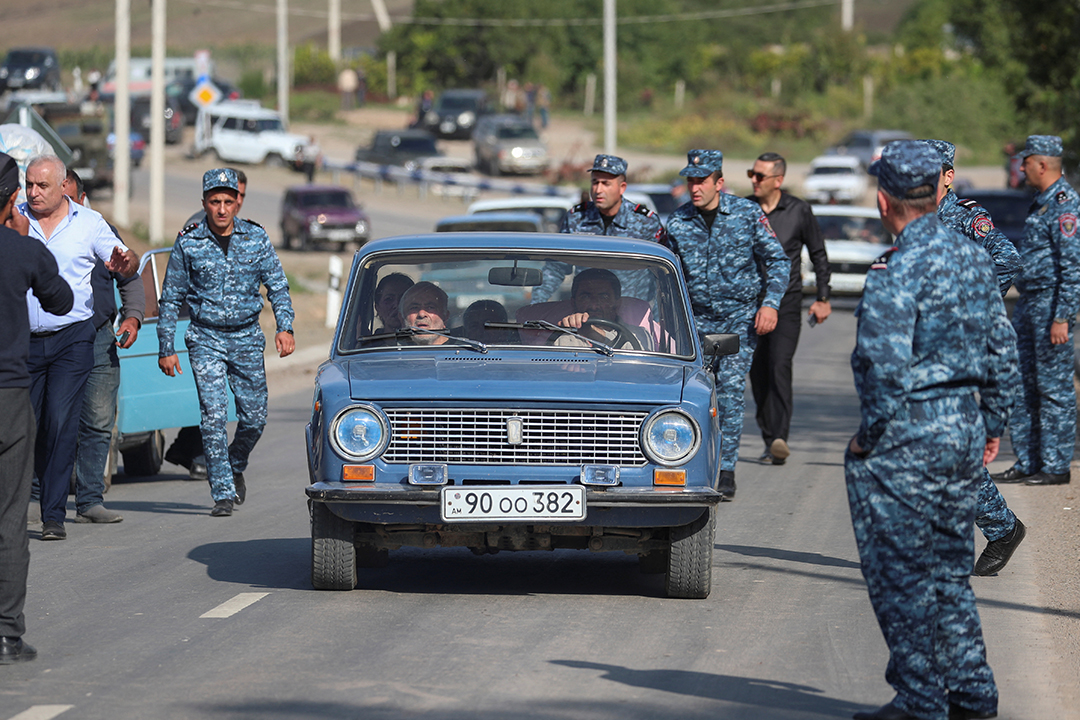 Soldiers in blue camouflage stand on a street as a blue car drives past.
