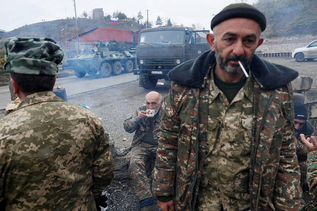 A soldier faces the camera smoking a cigarette while a military vehicle is in the background.