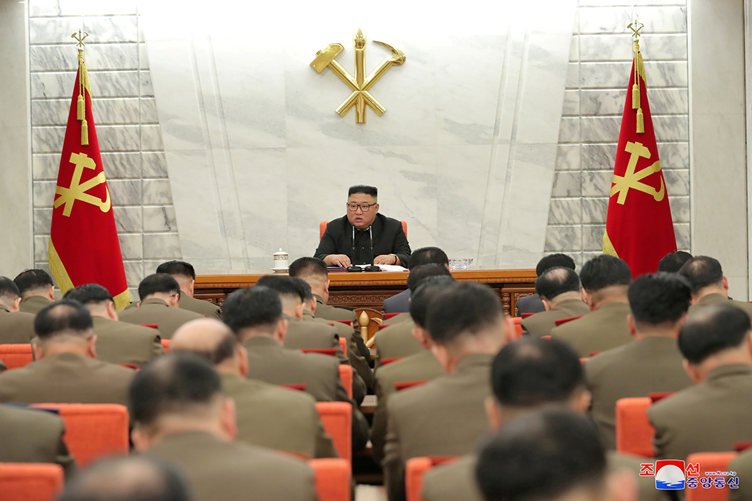 Kim Jong Un wears a white shirt and sits in front of a white wall with red and yelow flags while men sit facing him wearing tan jackets.