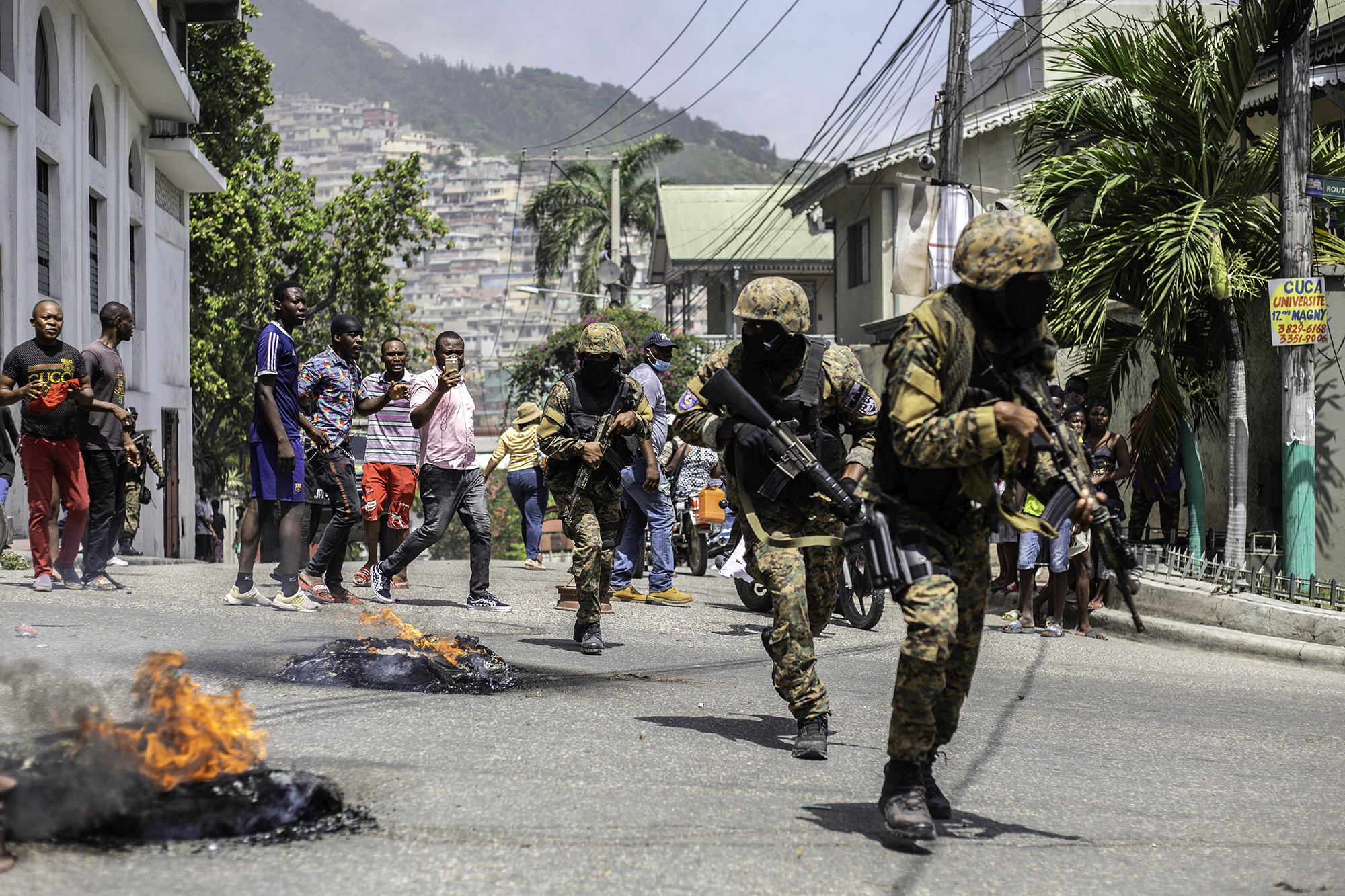 Soldiers with guns while civilians look on and tires burn in the street.