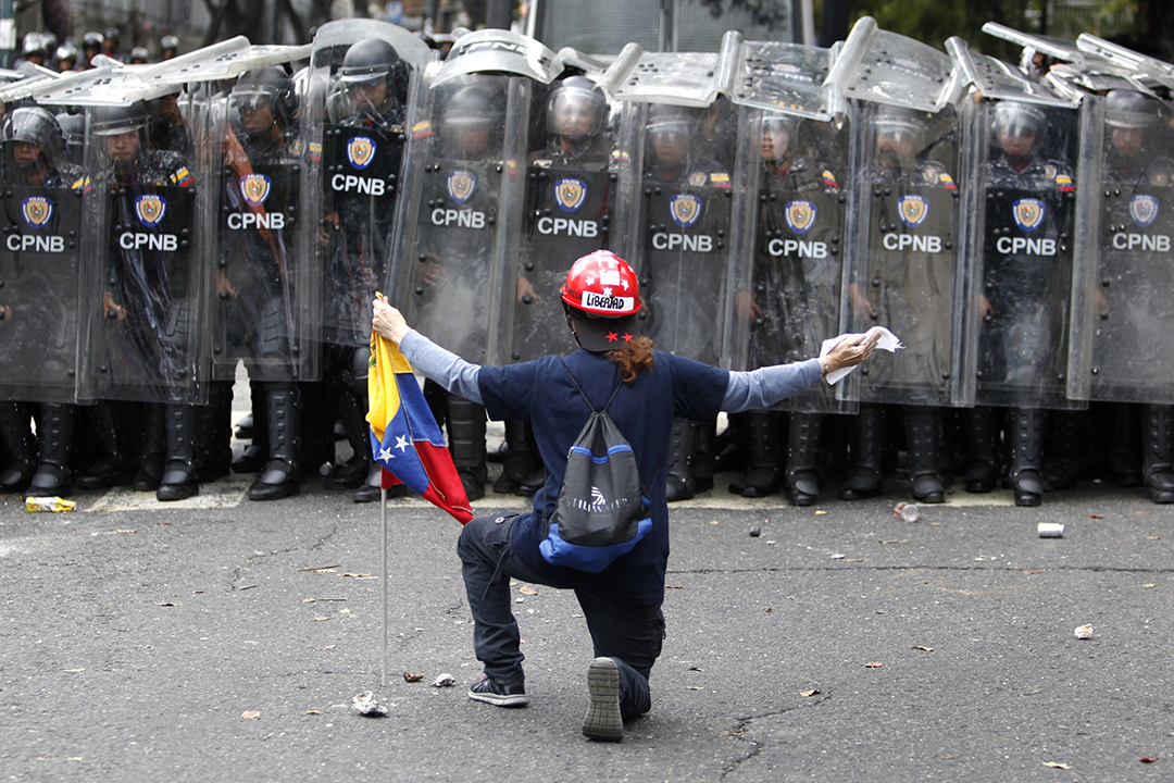 A man kneels facing police in riot gear.