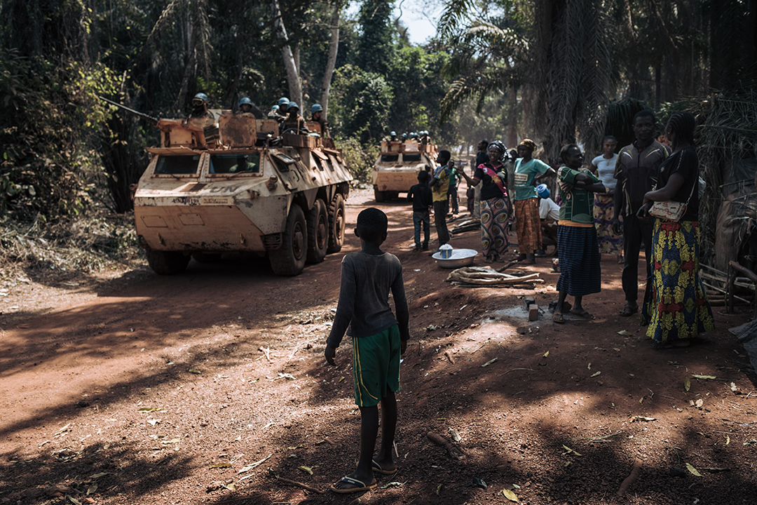 A boy stands facing a military tank in a forested area.
