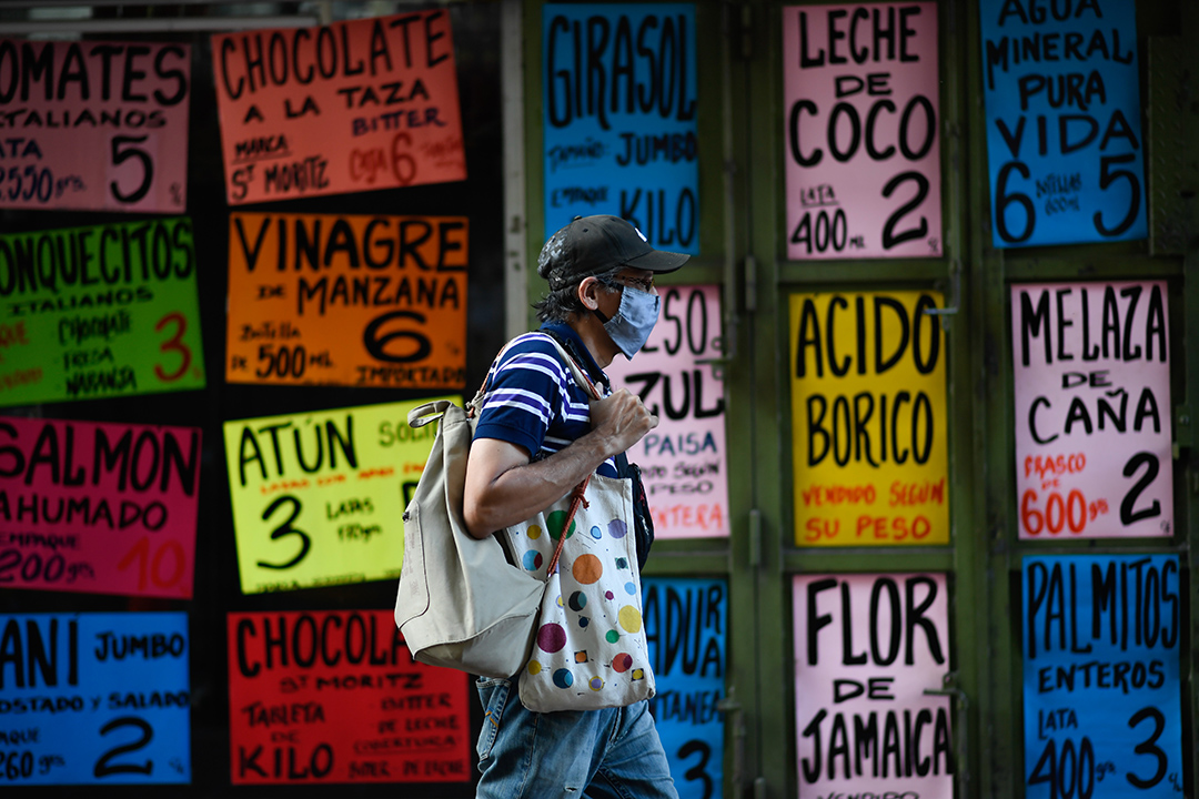 A man wearing a baseball hat and face mask walks past an array of colorful signs..