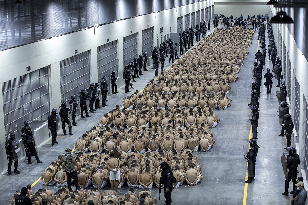 Two thousand men are seated and handcuffed on a prison floor facing away from the camera while flanked by police.