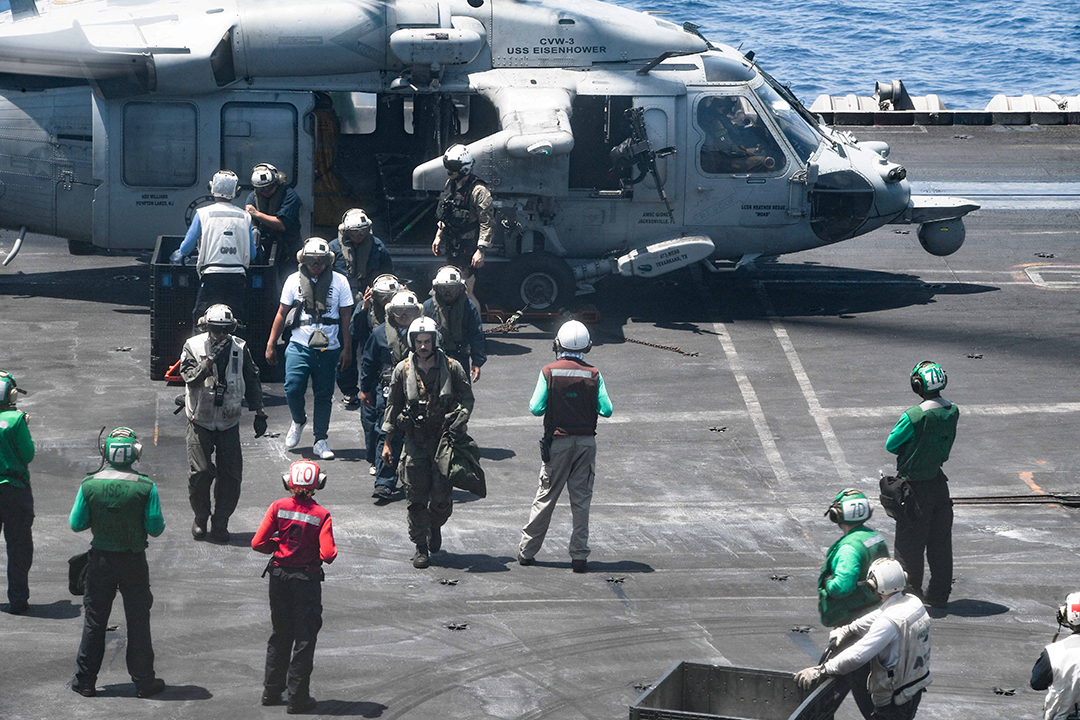 People wearing helmets walk off of a helicopter on a landing pad next to the sea.