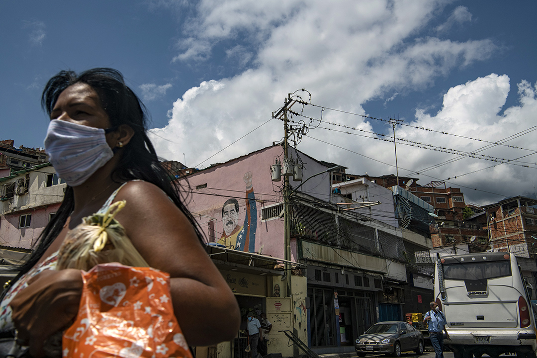 A woman wears a face mask walking away from a pink building with a mural of Nicolas Maduro.